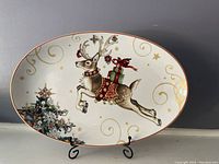 Oval ceramic platter with leaping reindeer and Christmas tree design on white background with gold swirls and stars, red rim.