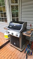 Full view of Weber Spirit BBQ with three burners and propane tank beside it on wooden deck floor. Rotisserie skewer visible extending from rear.