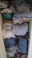 Multiple stacks of folded and rolled assorted bed linens including sheets, pillow cases, and blankets in neutral tones and blue.