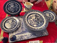 Set includes Blue Willow dinner and side plates, bowls, salt and pepper shakers, creamer and sugar bowl, and dinner mats as seen on red tablecloth.