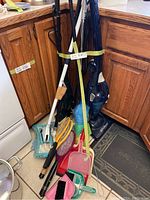 Photo showing the lot of cleaning tools grouped in a corner by kitchen cabinets including brooms, mops, dustpans, bug zappers, and handheld vacuum cleaners.