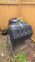 Photo showing both composters against a wooden fence, the rotating barrel style in front, secured with a white strap, and the vertical bin style behind it.