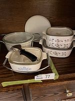 Full view of Corningware dishes and casseroles including square divided casserole dish with metal carrier, two rectangular casserole dishes with floral designs, glass round casserole dish with lid, and white round lid.