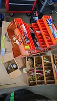 Two orange tackle boxes open showing assorted fishing lures and accessories placed inside, along with smaller boxes and fishing line containers.