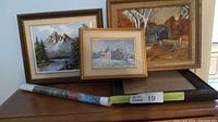 Three framed paintings including winter cabin, autumn covered bridge, and mountain lake landscape arranged on a table with two rolled posters underneath.