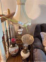 Three electric lantern style parlour lamps arranged together including one taller lamp with fabric shade, two smaller ceramic base lamps with metal frames and glass chimneys (two missing chimneys), plus one vintage patterned glass shade as bonus.