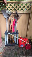Full view of long-handled garden tools arranged upright in a black plastic tool stand against an outdoor fence, including rakes, shovels, and red gas jugs on the ground.