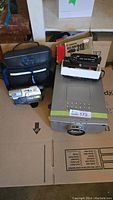 Overview of all items in lot: multimedia projector, Canon camera in box, Sony camcorder and carrying case on cardboard surface.
