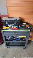 Front view of black steel rolling shop cart loaded with a black Husky toolbox, yellow handle tool, plastic sheets and hand tools.