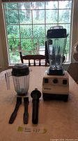 Front view of Vitamix base with large container attached, smaller container, tamper and two brushes laid out on table.