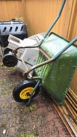 Green metal tub wheelbarrow with yellow-rimmed pneumatic tire, showing rust and wear on handles and tub.