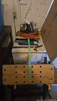 View showing one collapsible wooden workbench on metal frame with holes and measurement guides, circular saw placed on top with orange extension cords coiled beneath, and several metal prybars resting on the bench.