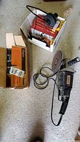 Overview of Motomaster battery charger in a box, Craftsman disk grinder on floor, and box of staples on concrete floor.