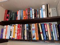 Bookshelf with two wooden shelves filled with a mix of hard and soft covered books including titles like 'The Last Good Year,' 'Living with History,' 'The Good German', and more.