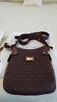 Front view of brown Coccinelle monogram crossbody purse with dark leather strap laying on white dust bag