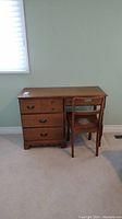 Wooden desk and chair placed in a room with light carpet and pale green walls. Desk has four drawers with metal handles and a medium brown finish.