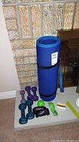 Blue rolled yoga mat with strap, various dumbbells, green stress ball, and black/pink wrist weights on white surface against brick fireplace.