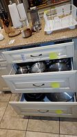 Kitchen countertop and open drawers showing stainless steel pots and pans, assorted kitchen utensils like a grater, and a white dish rack.