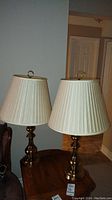 Two matching brass table lamps with cream pleated shades placed on a wooden table in a living room setting. Both lamps have brass bases and round brass finials on top of the shades.