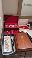 Photo showing wooden jewelry box open next to multiple jewelry pieces including pearl necklace, shell necklace, and various bracelets and watches.