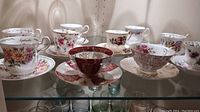 Overview of 7 assorted vintage teacups with matching saucers displayed on glass shelf showing various floral and gold designs