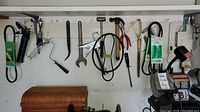 Complete view of hand tools hanging on pegboard including mallet, hammer, sanding belts, wrench, caulking gun, and paint roller