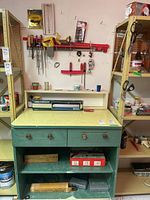 Photo of vintage green workbench with drawers and shelves holding toolboxes and supplies, tools mounted on the wall including clamp and screwdrivers.
