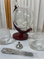 Full view showing globe-shaped decanter on wooden stand, two etched airplane whiskey glasses, metal funnel, and metal corkscrew with wood handle on white surface.