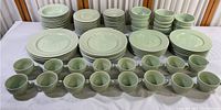 Full view of set of plates, bowls and cups arranged on white cloth. Shows large quantities of sage green ceramic dinnerware grouped by type.