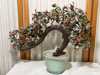 Overall view of the faux bonsai tree showing the curved textured trunk, pink flowers, green leaves, and green ceramic pot with white gravel filling.