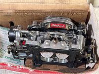 Top view of silver metallic Edelbrock carburetor inside a cardboard box showing throttle butterfly valves and mounting flange.