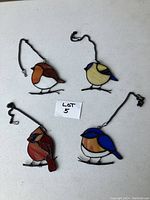 Four stained glass bird suncatchers displayed on a white surface with metal hanging chains visible. Various colors are seen including blue, orange, cream, red and brown.