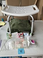 Photo showing white plastic adjustable shower stool with handles, green folded plush towels beneath, and various skincare and bath products including boxed Lever soap, Smith's hand cream, sample bottles and travel sizes around the stool base.