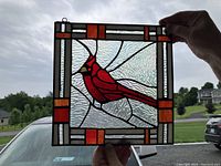 Stained glass panel held up to window showing red cardinal design with geometric red and orange border details.