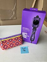 Purple boxed detangling hot air brush with colorful makeup pouch beside it on white surface.