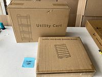 Box labeled 'Utility Cart' and 'HOPPEL PEG BOARD WITH HOOKS BLACK' showing unopened packaging for a rolling utility cart and peg board.