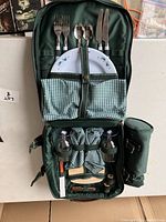Open picnic backpack showing tableware slots with forks, spoons, knives, and two plates secured inside. Also visible are fabric pouches and storage compartments, including bottle holders.