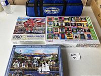 Three unopened 1000-piece puzzles titled Red Barn, Mediterranean Windows, and Jane Wooster Scott displayed on table.