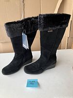 Full view of the pair of tall black suede boots with fold-over faux mink lining cuff and tags attached, standing upright.