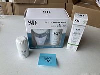 Boxed kit showing teeth whitening device with LED light, whitening gel bottle, and gum health product in clear window packaging alongside an extra whitening gel tube.