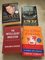 Four books neatly arranged on a gray surface: Money Master the Game by Tony Robbins, Think and Grow Rich by Napoleon Hill, The Intelligent Investor by Benjamin Graham, and The 4-Hour Workweek by Timothy Ferriss.