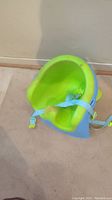 Green and blue Bumbo infant seat with safety straps, positioned on carpet against wall showing wear marks.