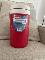 Red Coleman thermos jug on beige couch showing front label and white lid.