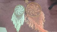 Two round crochet dream catchers laid flat on pink carpet, one off-white and one peach, with crochet patterns and long fringe tassels hanging down.