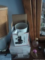 White Ember Mug partially inside an open box with product labels and markings, positioned on a table beside other household items.