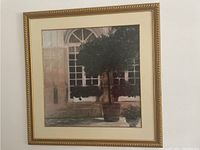 Framed print of a green potted tree sitting outside a building with large windows, with gold decorative frame and cream matting.
