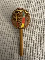 Front view of the wooden maraca showing painted geometric design in green, yellow, red, and white, with visible scratches and aged paint.