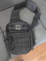 Front view of black backpack showing Canadian maple leaf patch, multiple zippered compartments, and adjustable padded shoulder strap.