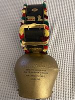 Image of brass cowbell showing the inscription and colorful tasseled leather strap with Swiss decorative plaques.