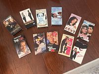 Top-down photo showing all 11 VHS romantic comedy movies arranged on wood floor, titles visible, including Stepmom, True Lies, Pretty Woman, Notting Hill, When Harry Met Sally, etc.
